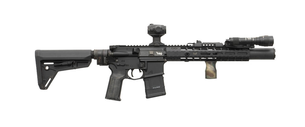 Photograph of a black home defense short-barreled rifle (SBR) displayed horizontally against a gray background with bold black text "HOME DEFENSE SBR" above. Rifle features adjustable stock, red dot sight, suppressor, flashlight, and vertical foregrip, highlighting tactical customization for close-quarters defense.