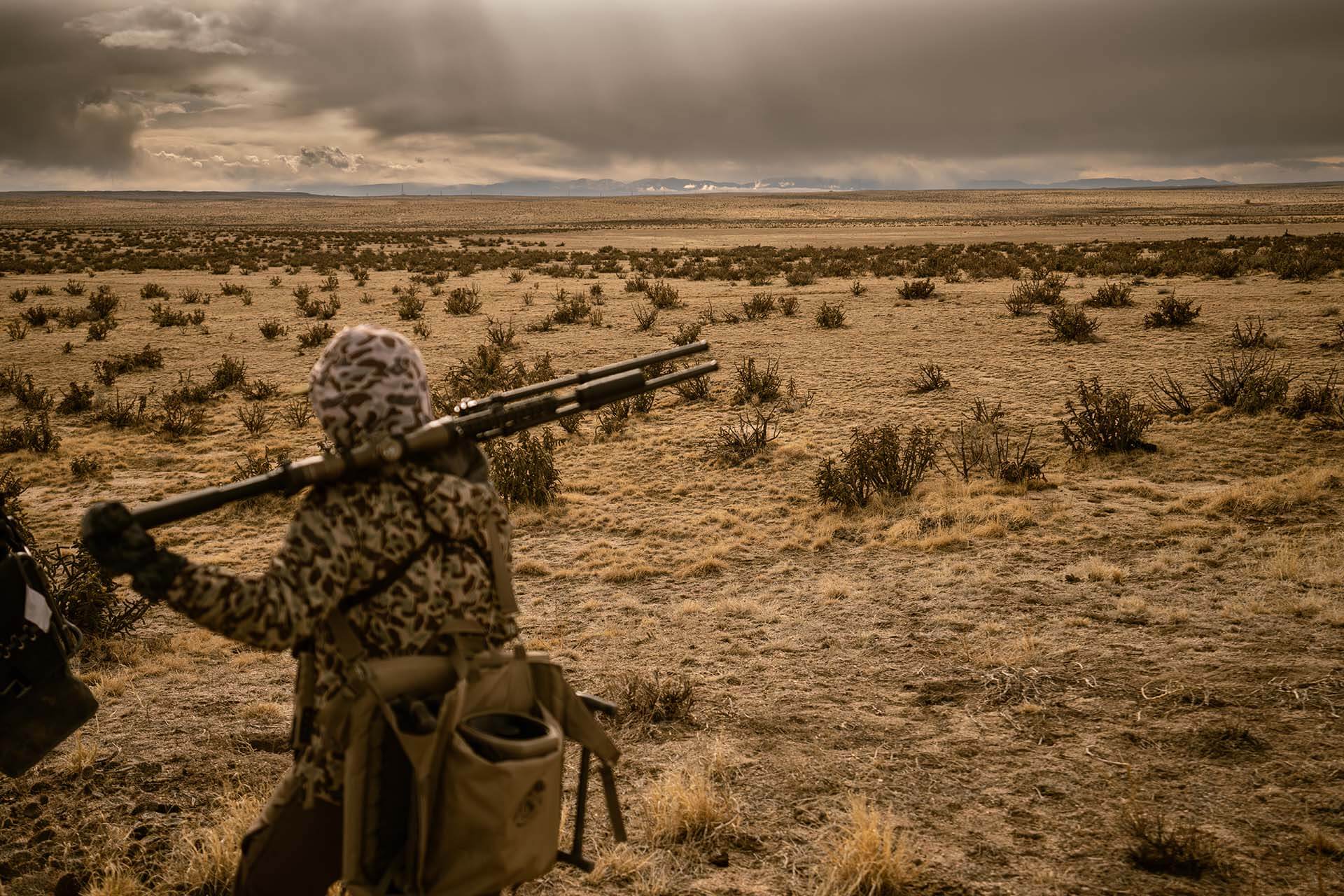 Hunter in full camouflage carrying a rifle across his shoulders while scanning an open desert landscape during a western coyote hunt.
