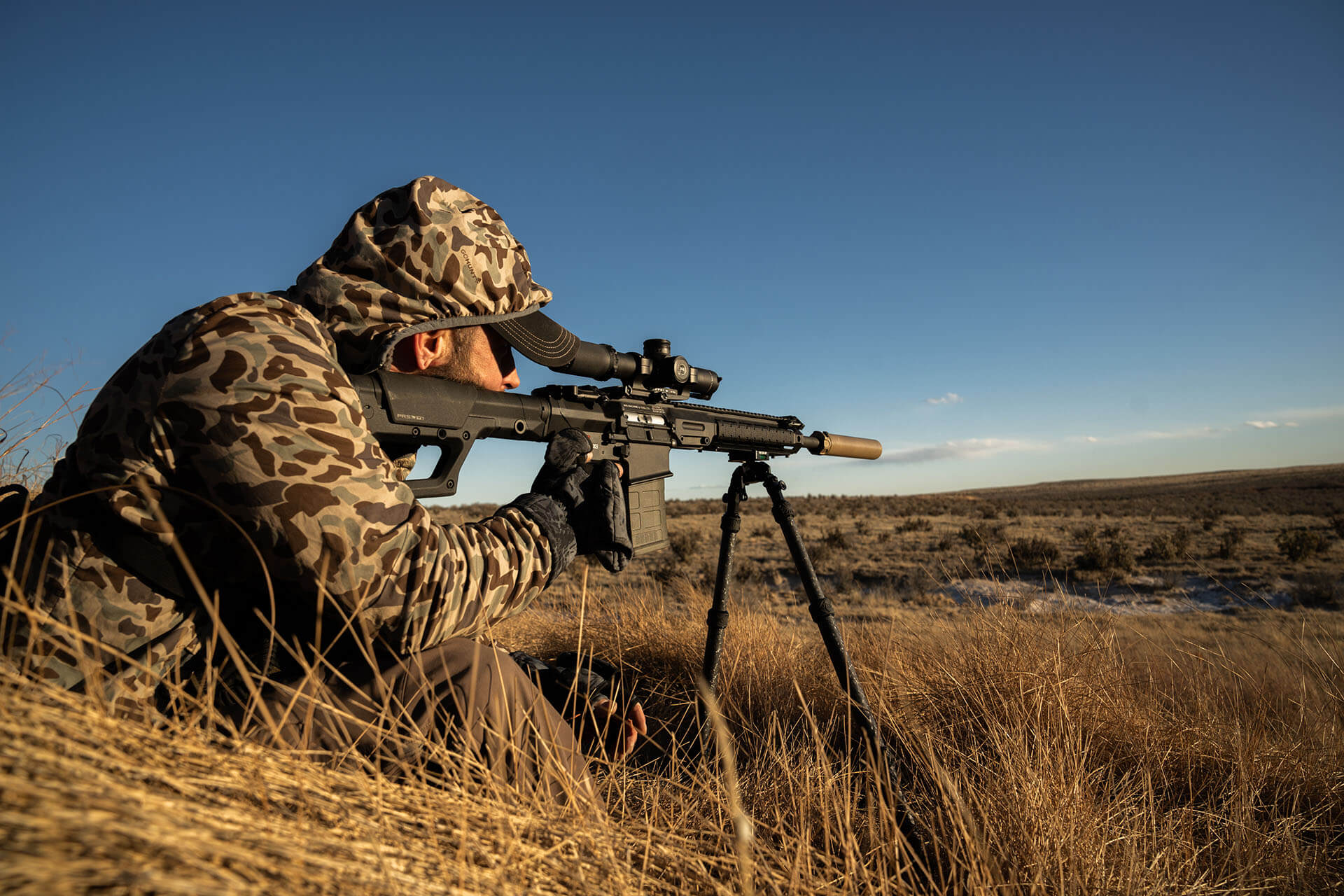 Profile view of a hunter in camouflage aiming an AR15 rifle equipped with a scope, suppressor, and bipod across a grassy field on a coyote hunt.