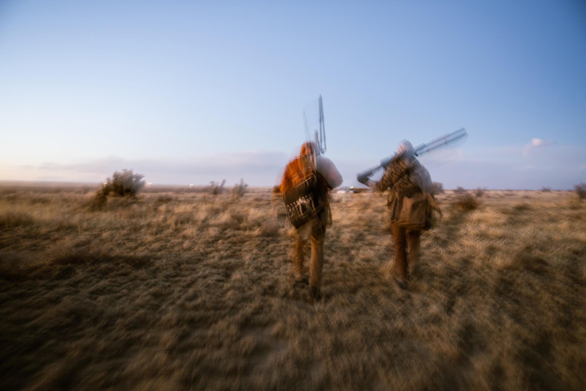 Motion blur shot of two hunters carrying rifles over their shoulders while walking through a grassy field at dawn.