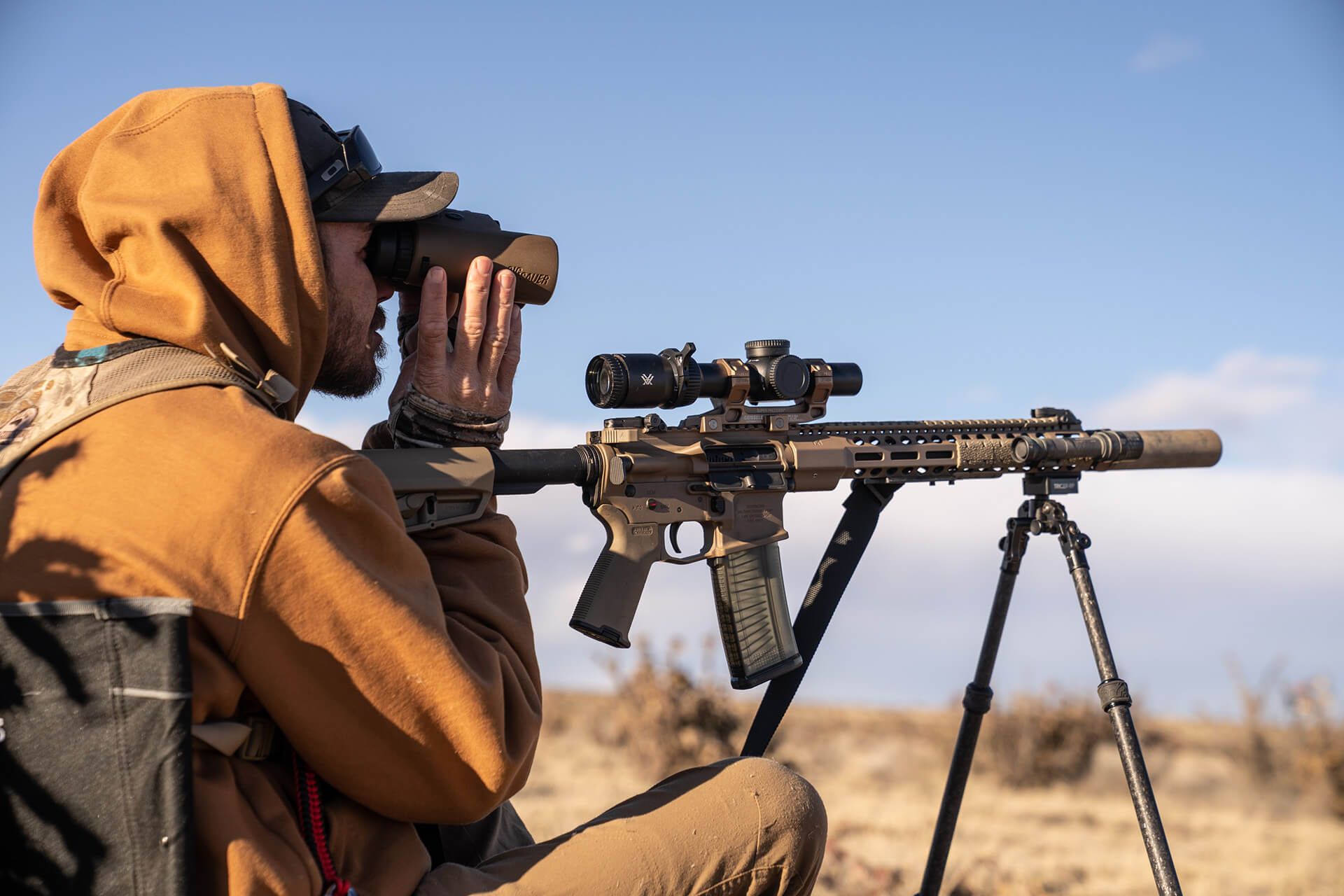 Side view of a hunter looking through binoculars next to a flat dark earth AR15 rifle mounted on a tripod setup during a coyote hunt.