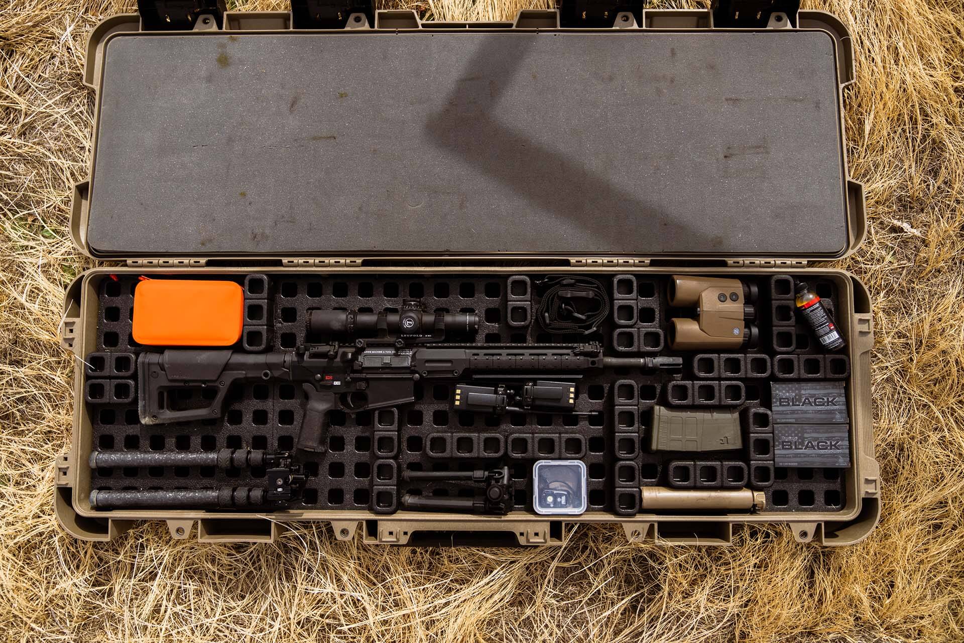 A black AR-pattern semi-auto rifle packed horizontally inside a tan modular hard case alongside binoculars, ammunition boxes, a suppressor, and a tripod on dry grass.